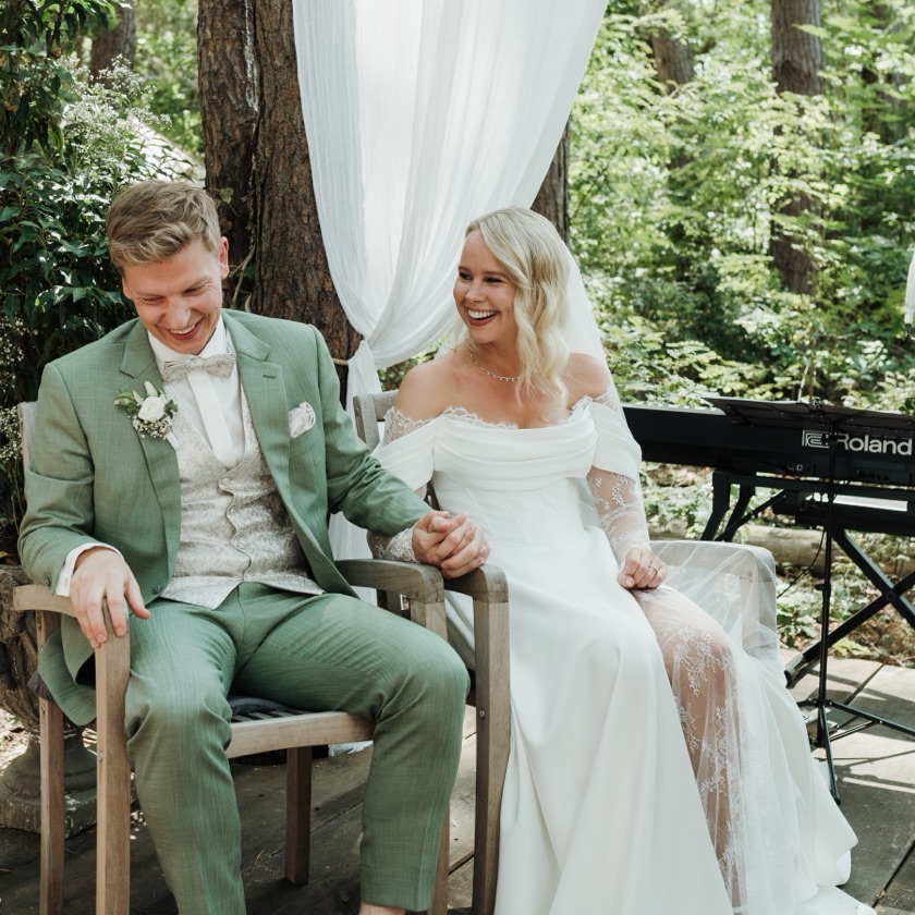 Viktoria und Christoph sitzen lachend Hand in Hand bei ihrer Trauung im Wald